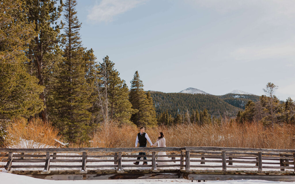 couple walking on bridge for Rocky Mountain National Park elopement surrounded by trees, mountains, and snow