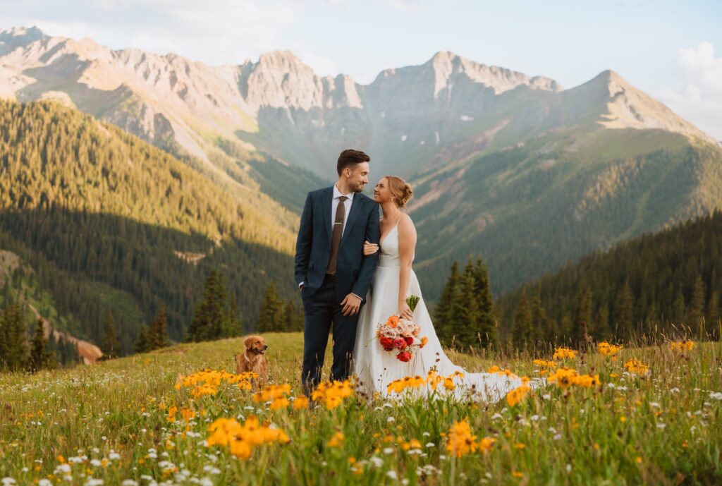 bride, groom, and dog posing for elopement pictures in field with flowers backed by mountains