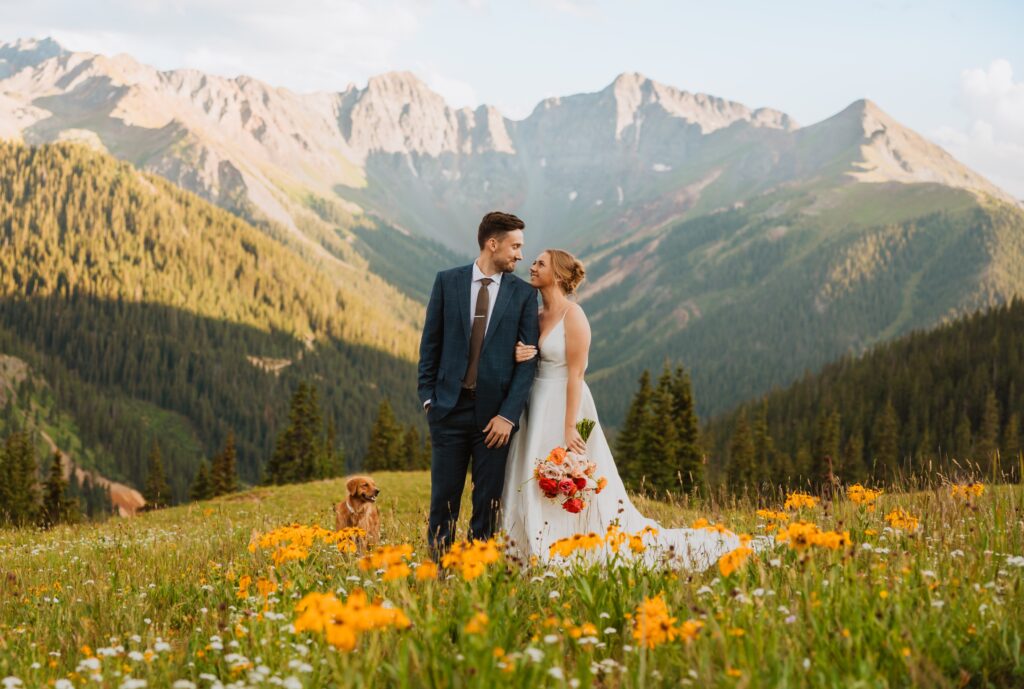 bride, groom, and dog in field with wildflowers