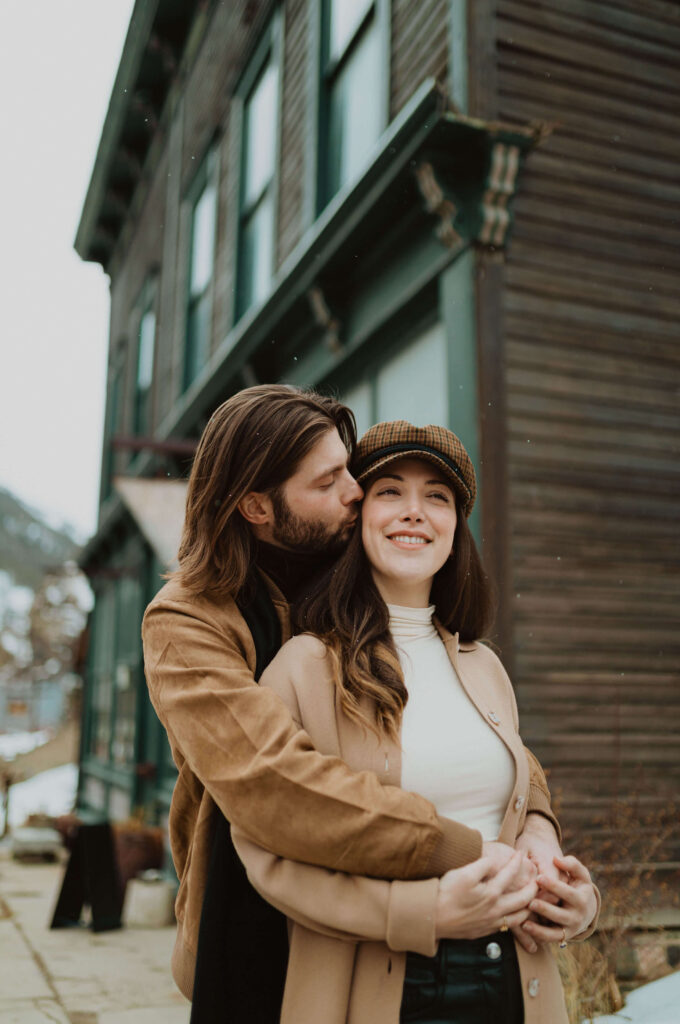 Couple wearing winter jackets and a hat standing outside in front of building posing for engagement photos