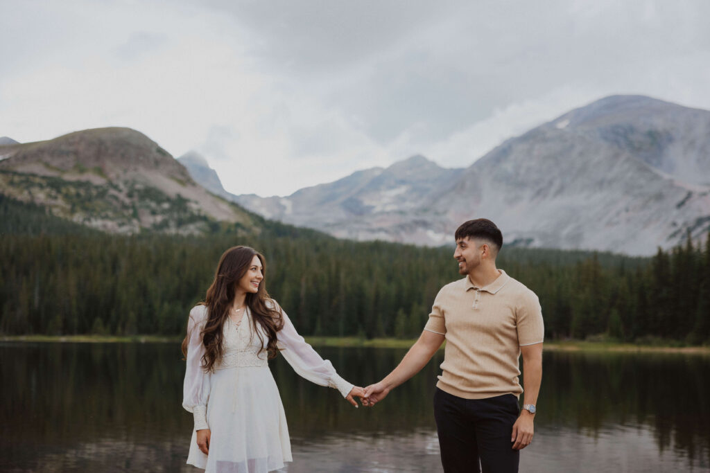 Woman wearing white dress and man wearing beige top and dark pants posing for engagement photos in front of a lake and mountains