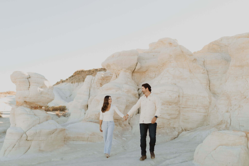 man and woman holding hands and wearing white tops and dark pants posing for engagement photos