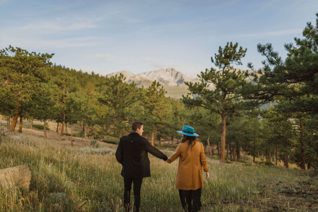 Couple walking in meadow towards trees and mountain scenery for Rocky Mountain National Park engagement photos