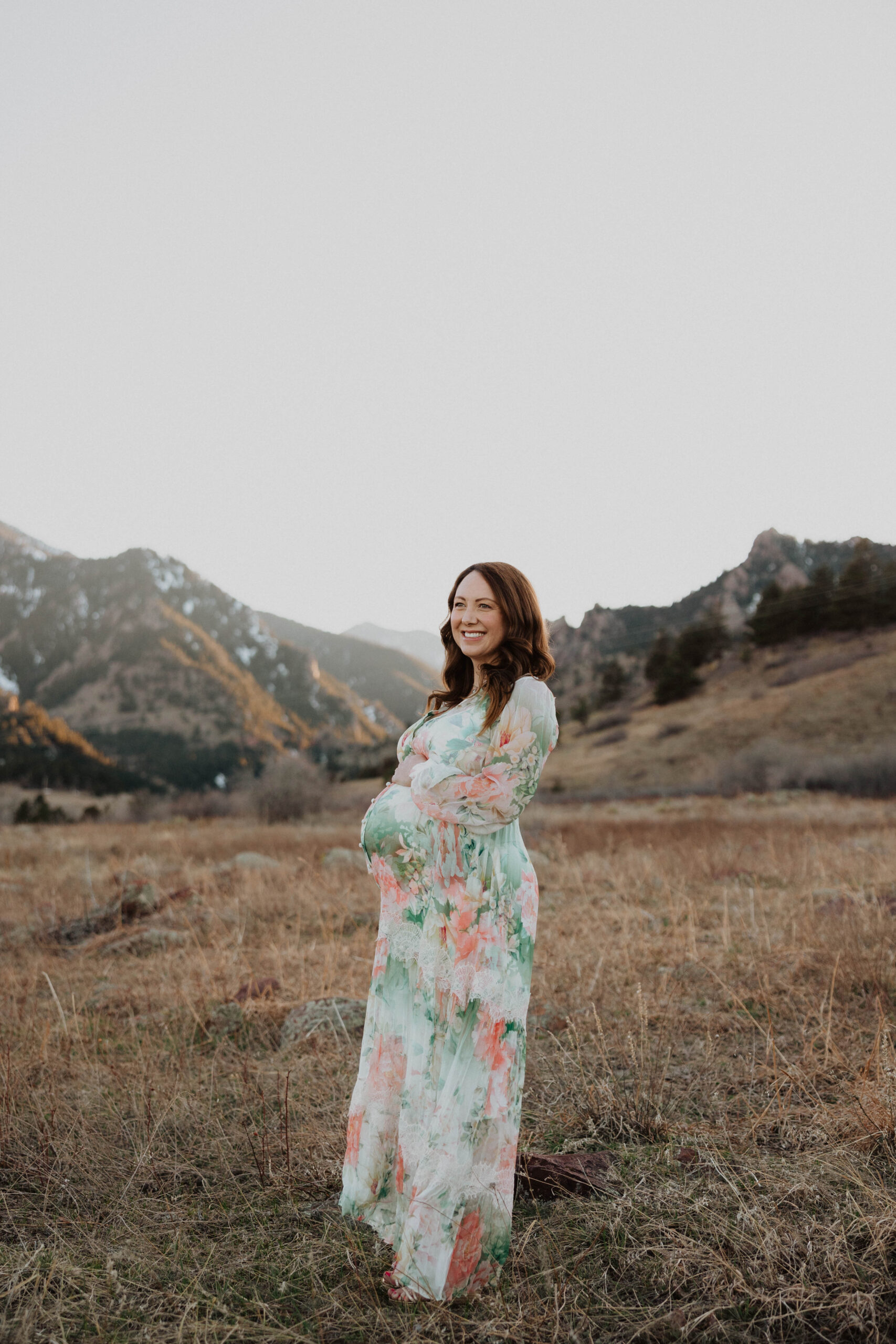 Pregnant mom holding stomach and smiling surrounded by mountain scenery in Denver