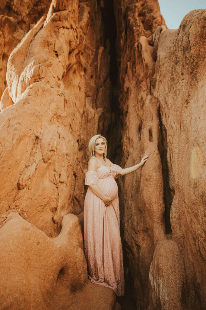 Woman standing in front of a red rock formation holding baby bump and smiling at camera