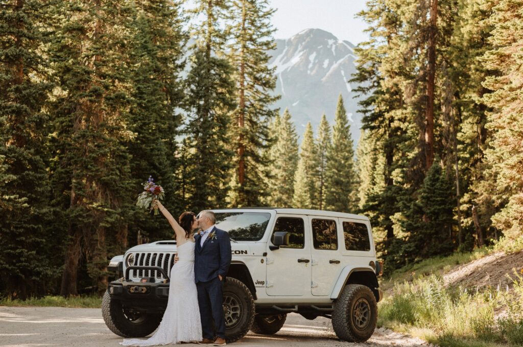 Bride and groom kissing next to white jeep surrounded by aspen trees and mountains