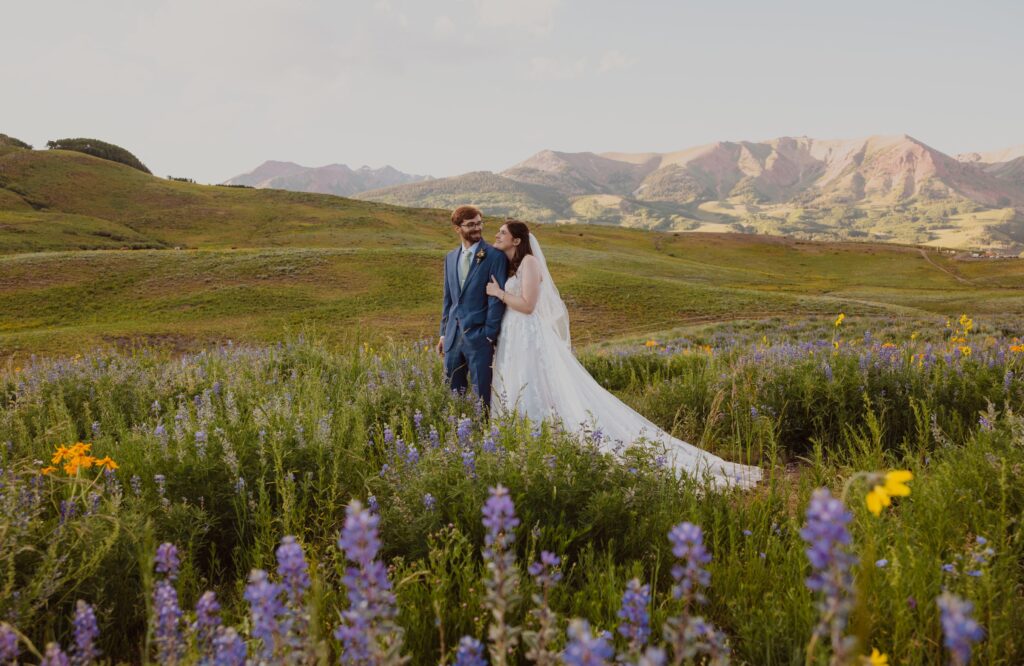 Bride and groom smiling at each other surrounded by flowers and mountain scenery