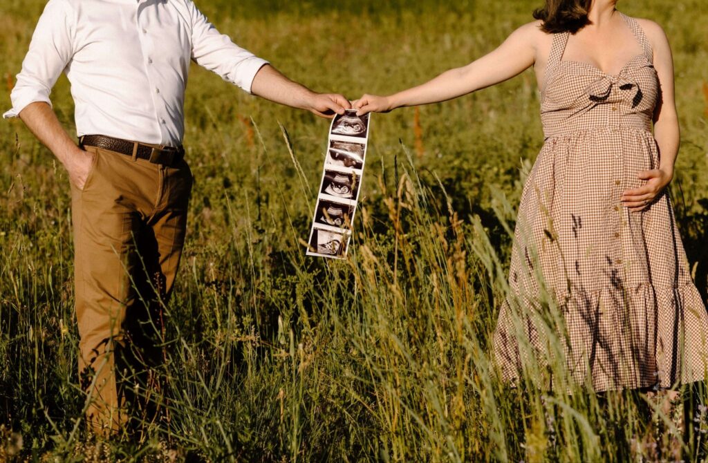 Man and pregnant woman posing in field and holding sonogram