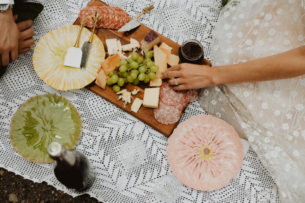 Overhead picture of a charcuterie board on picnic blanket with bride and groom in the periphery.
