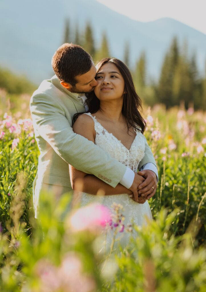 Groom embracing bride from behind while surrounded by pink wildflowers.