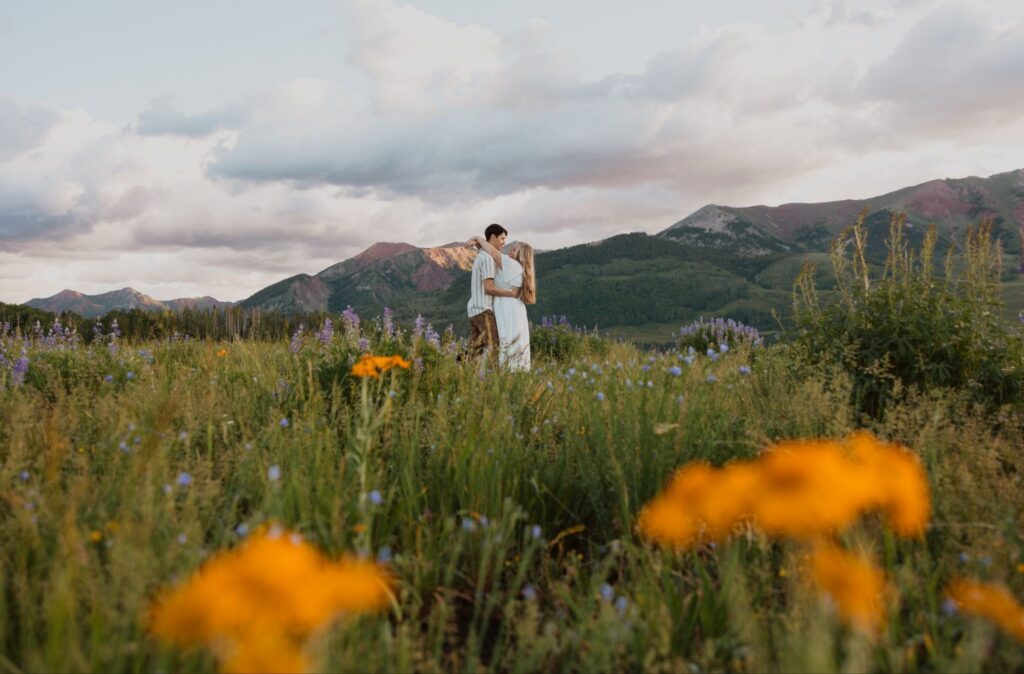 A couple embraces among wildflowers during a Crested Butte proposal with orange wildflowers in the foreground.