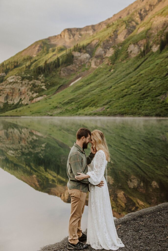 A couple standing beside water lean in for a kiss with mountains in the background.