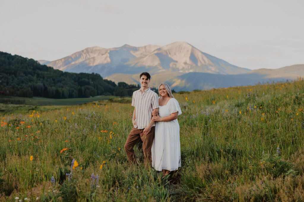 A couple poses among purple and orange wildflowers with mountains in the background.