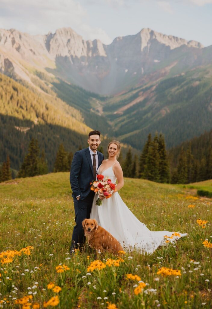 Bride and groom pose with their dog among wildflowers with mountains in background.