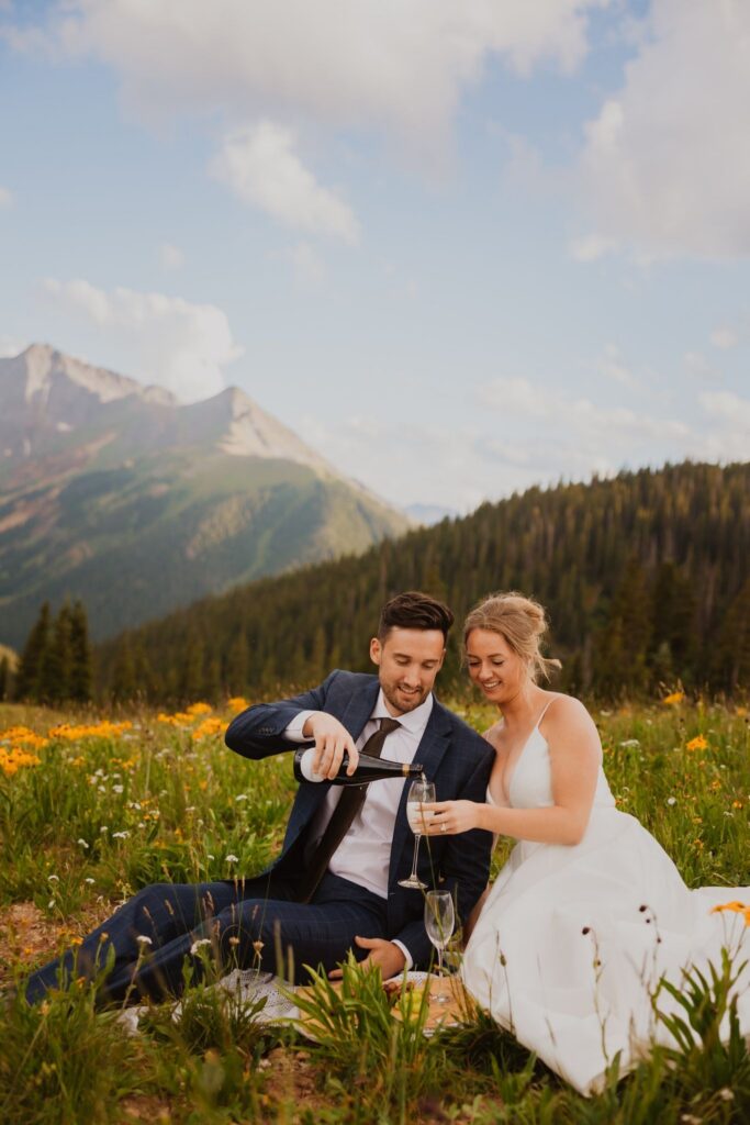 Groom pouring champagne for bride while they sit among wildflowers with pine trees and mountains in the background.