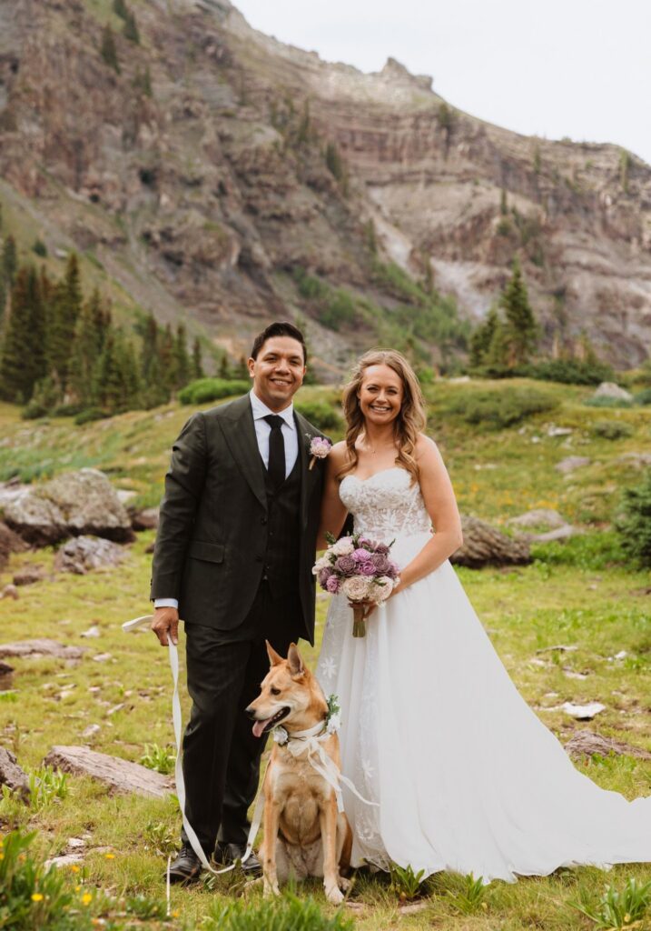 Bride and groom pose with their dog with mountains in the background.