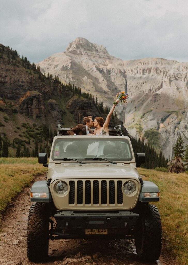 Bride and groom kiss in a Jeep with mountains and pine trees in the background.