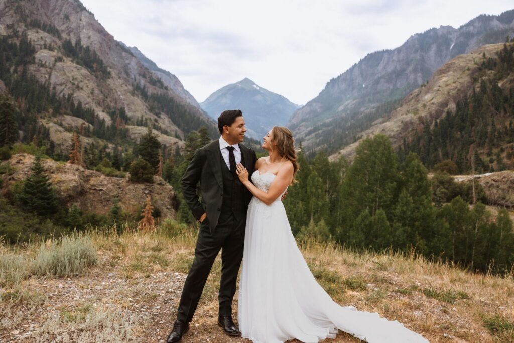 Bride and groom smile at each other with mountains and pine trees in the background.