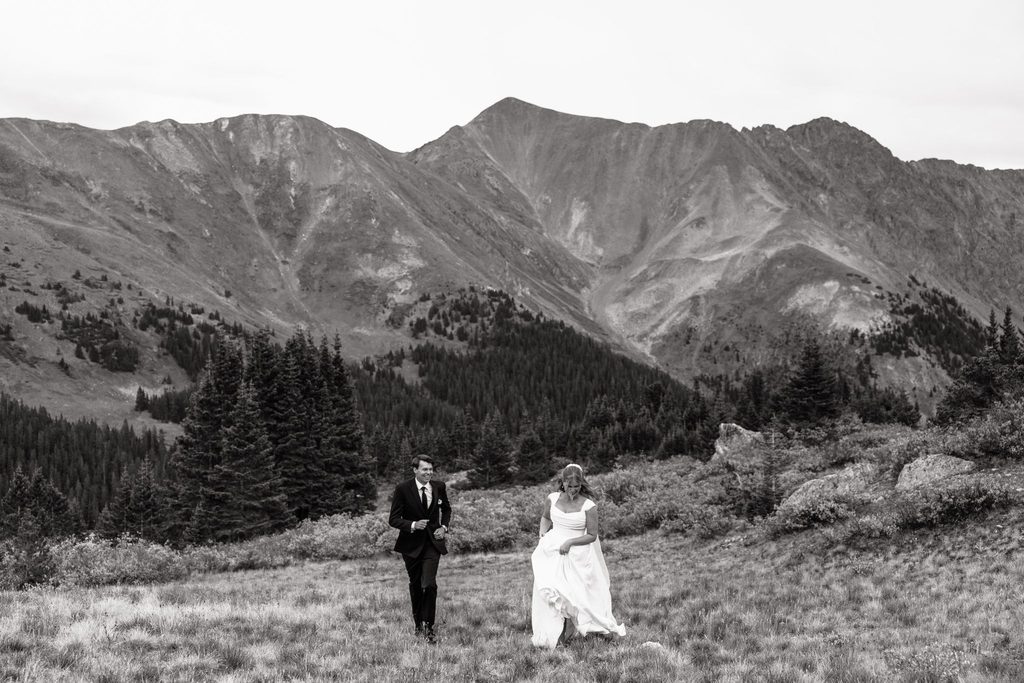 Black and white image of bride and groom running up hill with mountains and pine trees in the background.